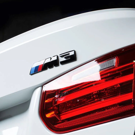 Gloss black BMW M3 replacement badge with blue, red, and white colour accents on the rear of a white BMW F80 M3 (Close Up)