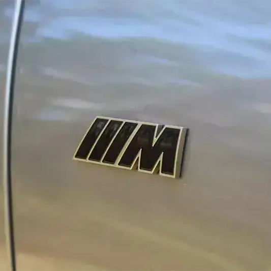 A pair of gloss black and gold side fender M badges for BMW G Series vehicles.